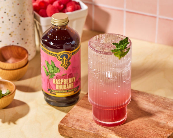 A tall glass with bright pink syrup fading into sparkling water sitting next to a bottle of Raspberry Rhubarb syrup on a kitchen counter.