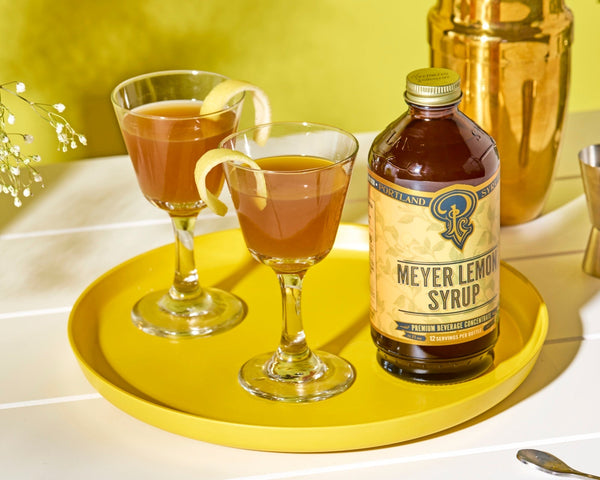 Two coupes each with a Oregon Gold Rush cocktail on a yellow tray with a bottle of Meyer Lemon syrup
