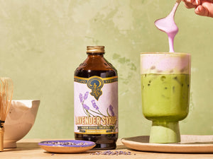 Bottle of Lavender Syrup with an iced matcha latte and lavender foam being poured.