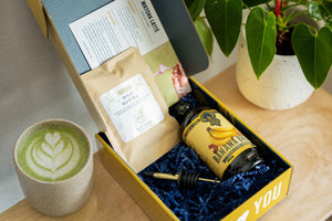 Matcha box open showing pouch of matcha, Banana Bread Syrup, pour spout, and recipe card. Matcha latte is next to the box.
